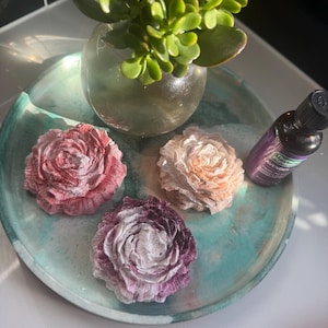 May include: Three decorative flower-shaped soap dishes in pink, peach, and purple shades on a green and blue tray with a small bottle of lavender essential oil.