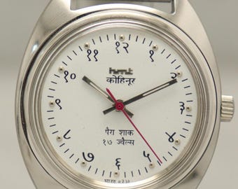 Puede incluir: Reloj de pulsera plateado con esfera blanca y correa plateada. La esfera del reloj tiene números negros y texto en idioma extranjero. Las manecillas del reloj son rojas. El reloj se exhibe sobre un soporte negro.