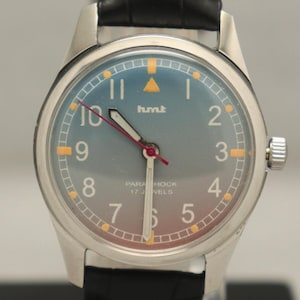 May include: A silver wristwatch with a blue and grey gradient dial, Arabic numerals, and the brand name "hmt." It has a black leather strap and a red second hand. The dial also reads "Parashock 17 Jewels."