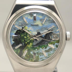 Puede incluir: Un reloj plateado con una correa de cuero negro. La esfera del reloj presenta una pintura de paisaje con cielo azul, árboles verdes y montañas. La fecha 14 es visible. El reloj tiene un bisel plateado y una corona.