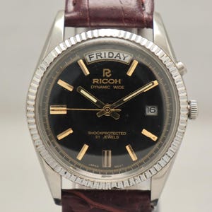 May include: A vintage Ricoh Dynamic Wide wristwatch with a black dial, gold markers, and a date window at the 3 o'clock position. The watch has a silver-tone case, a textured bezel, and a brown leather strap. The dial displays "FRIDAY" and "SHOCKPROTECTED 21 JEWELS".