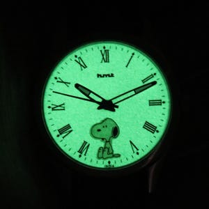 Puede incluir: Reloj que brilla en la oscuridad con esfera verde, manecillas negras y números romanos. El reloj presenta un diseño del personaje Snoopy. La palabra "HUMLE" está impresa encima del centro de la esfera. La palabra "INDIA" está impresa en la parte inferior.