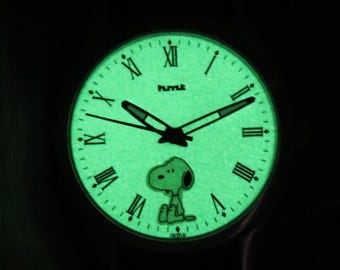 Puede incluir: Reloj que brilla en la oscuridad con esfera verde, manecillas negras y números romanos. El reloj presenta un diseño del personaje Snoopy. La palabra "HUMLE" está impresa encima del centro de la esfera. La palabra "INDIA" está impresa en la parte inferior.