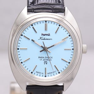 May include: A silver wristwatch with a light blue face and a black leather strap. The watch face displays the text "hmt Kohinoor" and "PARA SHOCK 17 JEWELS". The watch is marked "MADE IN INDIA 0231".
