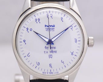 Puede incluir: Reloj de pulsera plateado con esfera blanca y números y agujas azules. El reloj tiene una correa de cuero negro y texto en idioma extranjero. El reloj se exhibe sobre un soporte transparente.