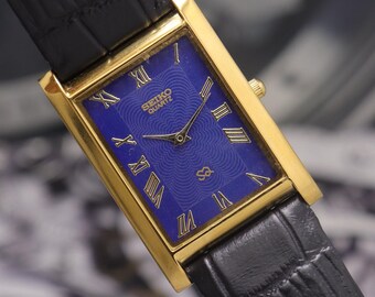 Puede incluir: Un reloj Seiko Quartz rectangular dorado con esfera azul y correa de cuero negro. La esfera del reloj presenta números romanos y el logotipo de Seiko Quartz. La correa es negra y texturizada.