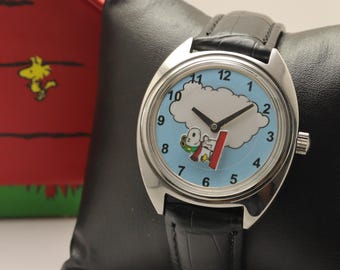 Puede incluir: Un reloj analógico plateado con esfera azul claro que presenta un gráfico de Snoopy, números negros y una correa de cuero negra. A su lado hay una caseta de perro de cartón roja con el nombre SNOOPY impreso en un letrero de madera.