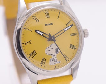 Puede incluir: Un reloj amarillo con caja plateada y correa de cuero amarilla. La esfera del reloj es amarilla con números romanos negros y un personaje de dibujos animados de Snoopy. El reloj tiene el texto "hmt" e "INDIA" en la esfera.