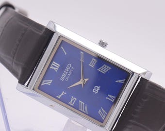 Puede incluir: Un reloj Seiko Quartz rectangular con esfera azul y caja plateada. El reloj presenta números romanos y una correa de cuero gris oscuro. El reloj se exhibe sobre un soporte transparente.