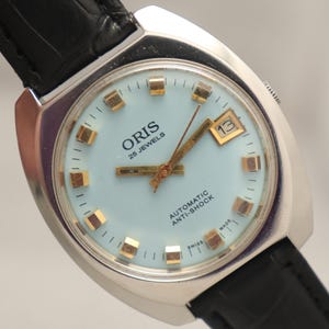 Peut inclure: Une montre Oris vintage avec un cadran bleu clair, des aiguilles et des index dorés, et un guichet de date. Le boîtier est argenté et le bracelet en cuir noir. Le cadran indique "ORIS 25 JEWELS AUTOMATIC ANTI-SHOCK SWISS MADE". Le bracelet mesure environ 20 cm de long.