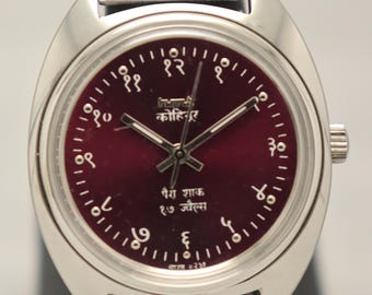 Puede incluir: Un reloj de pulsera plateado con esfera burdeos y correa plateada. La esfera del reloj presenta números arábigos y texto en idioma extranjero. El reloj se exhibe sobre un soporte negro.