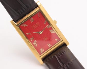 Puede incluir: Un reloj Seiko Quartz rectangular dorado con esfera roja brillante y números romanos. El reloj tiene una correa de cuero marrón con un patrón de cocodrilo. La esfera del reloj muestra el logotipo de Seiko Quartz.