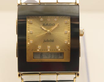 Puede incluir: Un reloj de pulsera Rado Jubilé con esfera cuadrada negra, dial analógico dorado, marcadores de hora de diamante y pantalla digital en la parte inferior. La correa tiene eslabones metálicos plateados con forma de pirámide.