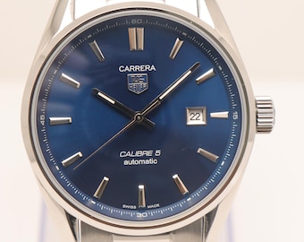 Puede incluir: Un reloj TAG Heuer Carrera de acero inoxidable con esfera azul profundo, manecillas e índices plateados, una ventana de fecha en la posición de las tres y el texto Carrera, TAG Heuer, Calibre 5 automatic y Swiss Made en la esfera.