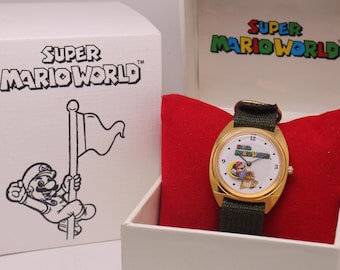 Puede incluir: Un reloj de pulsera dorado con esfera blanca que presenta gráficos de Super Mario World, una correa de tela verde oscuro y un cojín de terciopelo rojo dentro de una caja de presentación blanca. La tapa y el exterior muestran el logo de Super Mario.