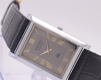 Puede incluir: Un reloj Seiko Quartz rectangular con caja plateada y esfera gris con números romanos dorados. El reloj tiene una correa de cuero negro con un patrón de cocodrilo. El reloj está en ángulo, mostrando su diseño.