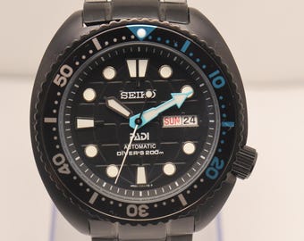Puede incluir: Un reloj de buceo automático Seiko PADI negro con brazalete de eslabones de acero inoxidable. La esfera negra presenta marcadores de hora luminosos, una ventana de día y fecha a las tres, un minutero azul y un bisel giratorio.