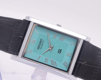 Puede incluir: Un reloj Seiko Quartz rectangular con caja plateada y esfera turquesa. El reloj presenta números romanos y una correa de cuero negro. La esfera del reloj tiene el logotipo de Seiko y un diseño estilizado. La correa tiene un patrón de cocodrilo.