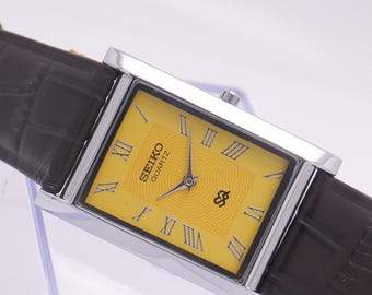 Puede incluir: Un reloj Seiko Quartz rectangular con caja plateada y correa de cuero marrón. La esfera del reloj es amarilla con números romanos y agujas negras. El reloj tiene el logotipo de Seiko Quartz.