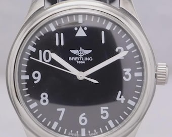 Puede incluir: Un reloj Breitling de acero inoxidable con esfera negra y números blancos. El reloj tiene una correa de tela gris y una corona plateada. La esfera muestra el logotipo de Breitling y el texto "1884".