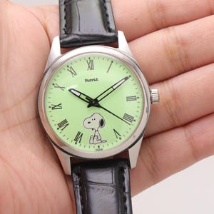 May include: A silver-toned wristwatch with a light green face featuring Snoopy. The watch has black hands, Roman numeral hour markers, and a black leather band. The watch face also has the word "INDIA" printed on it.