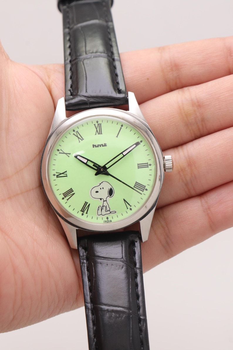 May include: A silver-toned wristwatch with a light green face featuring Snoopy. The watch has black hands, Roman numeral hour markers, and a black leather band. The watch face also has the word "INDIA" printed on it.