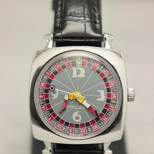 May include: A silver wristwatch with a black leather strap. The watch face has a roulette wheel design with red, black, and green segments. The hands are yellow and red. The watch reads "SHOCK PROC 17 JEWELS".