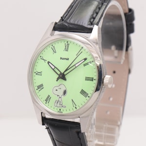 May include: Silver-tone wristwatch with a light green face featuring Snoopy. The watch has black Roman numerals, hands, and a black leather band. The watch face also has the text "hmt" and "INDIA".