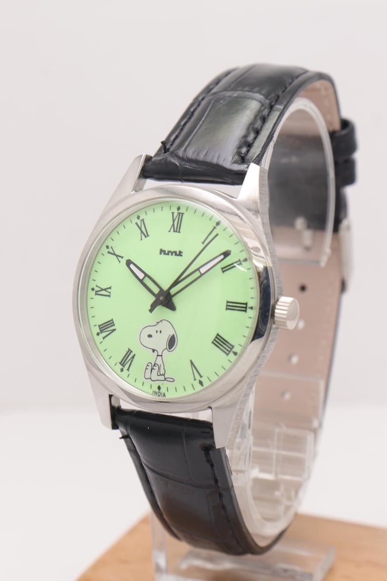 May include: Silver-tone wristwatch with a light green face featuring Snoopy. The watch has black Roman numerals, hands, and a black leather band. The watch face also has the text "hmt" and "INDIA".
