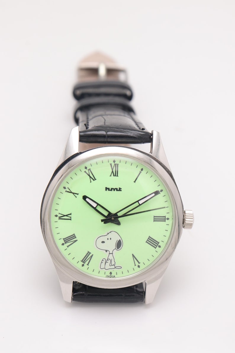 May include: Silver-tone wristwatch with a light green face featuring Snoopy and Roman numerals. The watch has black hands and a black leather band. The word "hmt" is printed on the watch face.