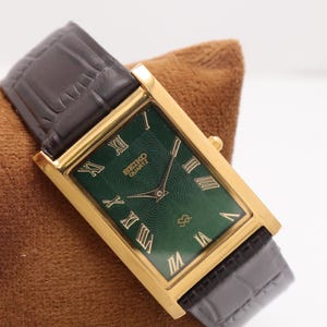 May include: A gold-toned rectangular Seiko Quartz watch with a dark green face and Roman numeral hour markers. The watch has a dark brown leather band. The watch is displayed on a brown surface.