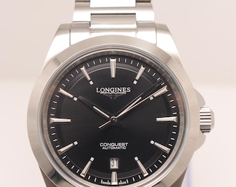 Puede incluir: Un reloj Longines Conquest Automatic de acero inoxidable con esfera negra, manecillas e índices plateados, una ventana de fecha en la posición de las seis y una pulsera de eslabones de metal a juego con cierre desplegable seguro.