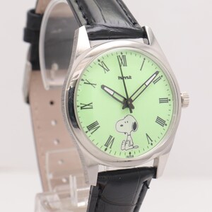 May include: A silver-toned wristwatch with a light green face featuring Snoopy. The watch has black hands and Roman numeral hour markers. It has a black leather band and the word "INDIA" is printed on the watch face.