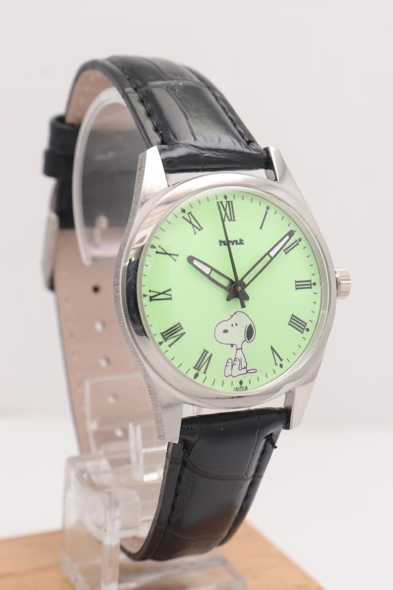 May include: A silver-toned wristwatch with a light green face featuring Snoopy. The watch has black hands and Roman numeral hour markers. It has a black leather band and the word "INDIA" is printed on the watch face.