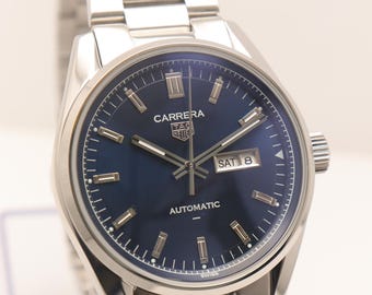 Puede incluir: Un reloj de pulsera de acero inoxidable plateado con esfera azul oscuro, que incluye el texto CARRERA, TAG HEUER y AUTOMATIC. El reloj cuenta con marcadores de hora plateados, una ventana de día y fecha que muestra SAT 8 y brazalete de eslabones.