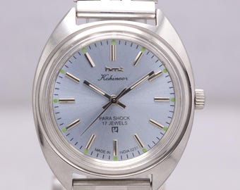 Peut inclure: Montre-bracelet argentée avec un cadran bleu clair. La montre affiche le texte "hmt Kohinoor" et "PARA SHOCK 17 JEWELS". Le bracelet est composé de maillons argentés. La montre mesure environ 3,8 cm de diamètre.