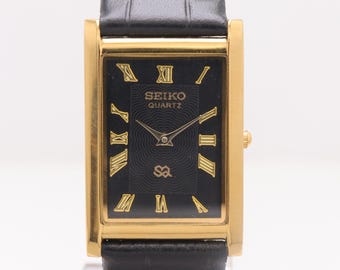 Puede incluir: Un reloj Seiko Quartz rectangular con esfera negra y números romanos dorados. El reloj tiene una caja dorada y una correa de cuero negro. El reloj se exhibe sobre un soporte transparente.