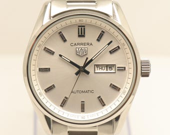 Puede incluir: Un reloj automático TAG Heuer Carrera de tono plateado con esfera plateada metálica, índices de bastón plateados y una ventana de día y fecha en la posición de las tres. El reloj cuenta con una pulsera de eslabones de acero inoxidable.