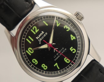 Puede incluir: Un reloj de pulsera HMT Jawan vintage con esfera negra y números verdes luminosos. El reloj presenta una caja plateada, una correa de cuero negro y un segundero rojo. La esfera del reloj incluye el texto "hmt Jawan" y "17 Jewels Para Shock".
