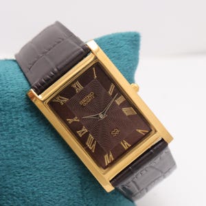 Puede incluir: Un reloj Seiko Quartz dorado rectangular con esfera marrón y números romanos. La correa es de cuero marrón con diseño de cocodrilo. El reloj se muestra sobre una superficie turquesa.