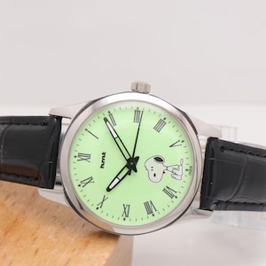 May include: A silver-toned wristwatch with a light green face featuring black Roman numerals and a Snoopy graphic. The watch has black hands and a black leather band. The watch face also has the text "hmt" and "India".