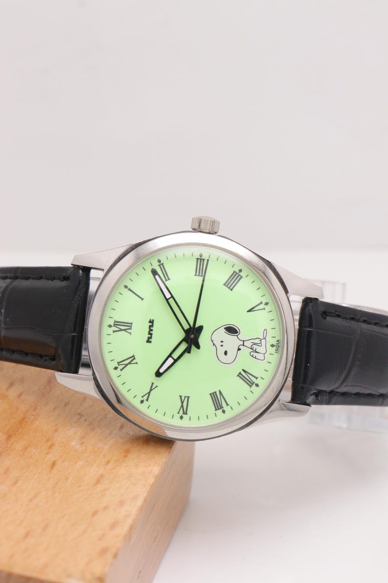 May include: A silver-toned wristwatch with a light green face featuring black Roman numerals and a Snoopy graphic. The watch has black hands and a black leather band. The watch face also has the text "hmt" and "India".