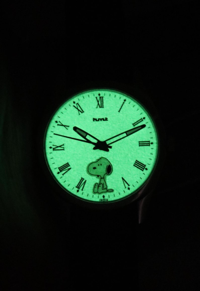 May include: Glow-in-the-dark watch with a green face, black hands, and Roman numerals. The watch features a Snoopy character design. The word "HUMLE" is printed above the center of the watch face. The word "INDIA" is printed at the bottom.