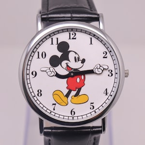 Pode incluir: Um relógio de pulso com caixa redonda prateada e mostrador branco com um gráfico do Mickey Mouse. Os braços da personagem servem como ponteiros das horas e minutos. O relógio tem algarismos arábicos pretos e uma pulseira de couro preta.