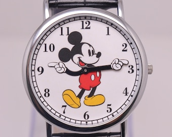 Puede incluir: Un reloj de pulsera con caja redonda plateada y esfera blanca que presenta un gráfico de Mickey Mouse. Los brazos del personaje sirven como manecillas de hora y minutos. El reloj tiene números arábigos negros y una correa de cuero negra.