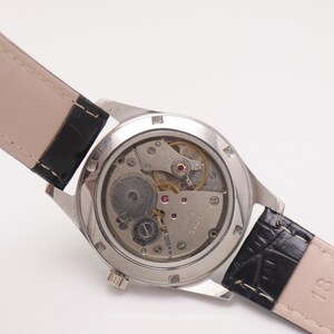 May include: A silver-toned wristwatch with a clear back, showcasing the intricate mechanical movement. The watch has a black leather strap with a textured pattern. The watch face is not visible.