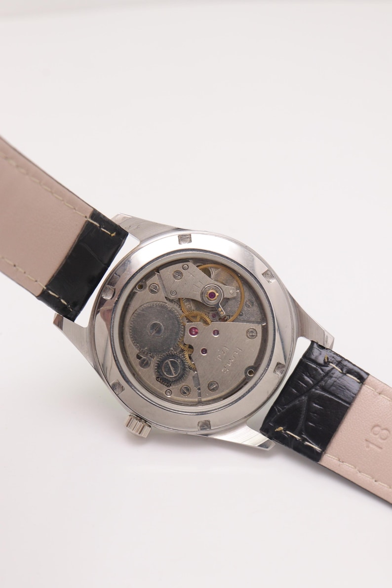 May include: A silver-toned wristwatch with a clear back, showcasing the intricate mechanical movement. The watch has a black leather strap with a textured pattern. The watch face is not visible.