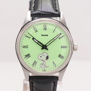 May include: A silver wristwatch with a light green face featuring Snoopy and Roman numerals. The watch has black hands and a black leather band. The word "hmt" is printed on the watch face, and the word "INDIA" is printed at the bottom.