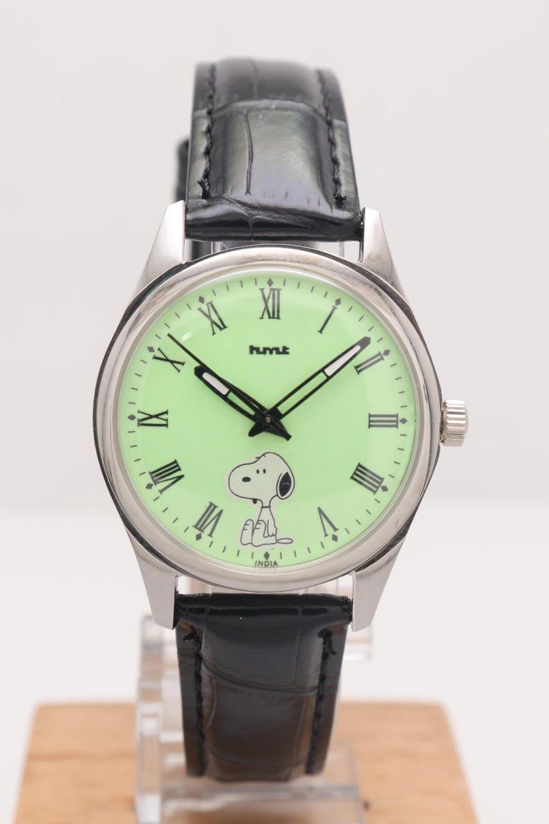 May include: A silver wristwatch with a light green face featuring Snoopy and Roman numerals. The watch has black hands and a black leather band. The word "hmt" is printed on the watch face, and the word "INDIA" is printed at the bottom.