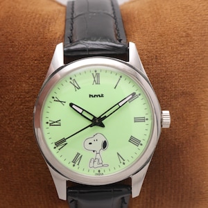 May include: A silver-toned wristwatch with a light green face featuring Snoopy. The watch has black hands and Roman numeral hour markers. The black leather band complements the watch face. The word "INDIA" is printed on the watch face.